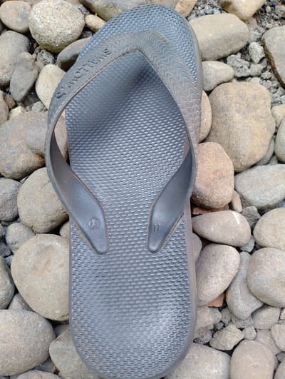 A dark gray flip-flop is laid on a bed of smooth, gray, and brown river stones. The flip-flop has a textured surface with a pattern of small raised ovals and a wavy pattern along the edges. The brand name "SWALLOW" is embossed on the strap. A small symbol resembling a peace sign is present on one of the straps, and the number "11" is visible on the other. The lighting suggests it is daytime, with soft shadows indicating an overcast sky or diffused sunlight. The environment appears to be outdoors, possibly near a river or in a rocky area. No people or animals are present in the image. The scene is static and unposed.