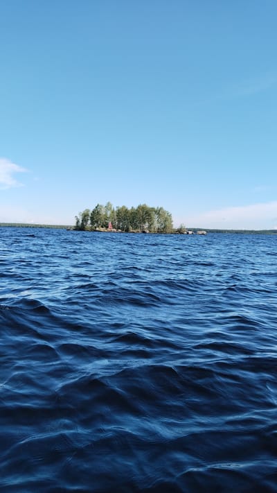 The image depicts a serene body of water, likely a lake, with a small, tree-covered island in the distance. The island is sparsely populated with trees, and a small red structure, possibly a lighthouse or a small building, is visible among them. The water is a deep blue with gentle ripples, suggesting a calm day. The sky is a clear, bright blue, with a few wisps of clouds on the horizon. The overall scene evokes a sense of tranquility and natural beauty. This scene is likely located on one of the many lakes surrounding Nadvoitsy, Russia, a town known for its numerous waterways and natural landscapes.
