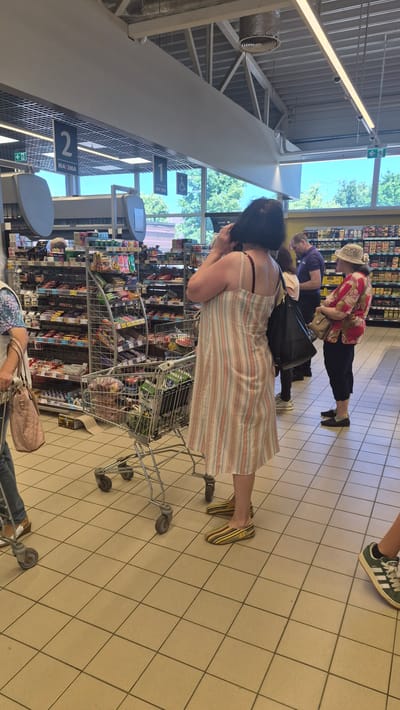 The image depicts a retail environment, likely a grocery store, in Preiļi, Latvia. The scene is viewed from a slightly elevated perspective, with the camera angled downwards. Several individuals are present, navigating the aisles with shopping carts.
In the foreground, a woman wearing a striped dress and yellow sandals is pushing a shopping cart. She is facing away from the camera, looking towards a display of products. Her posture suggests she is either browsing or selecting items.
Further into the background, other shoppers are visible. One person, wearing a floral print shirt and a hat, is also looking at merchandise. Another individual is seen from the back, wearing a purple shirt. The presence of multiple shoppers and stocked shelves indicates the store is operational and open for business.
The setting is characterized by tiled flooring and overhead lighting. Shelving units filled with various products are prominent, displaying a wide array of packaged goods. Overhead signs with numbers and text are visible, possibly indicating aisle numbers or product categories. One sign clearly reads "MAXIMA". The overall atmosphere appears to be that of a typical shopping trip. The lighting suggests it is daytime, and the presence of trees visible through a large window implies the store is situated in an area with natural light access. No specific emotions are clearly discernible from the visible subjects.