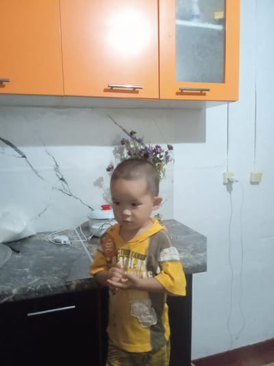 A young child stands in a kitchen, holding a lollipop. The child is wearing a yellow and brown long-sleeved shirt with some visible text on it. The kitchen features orange cabinets, a marbled countertop, and a dark cabinet below. There is a small white pot with a red lid on the counter, and a bag of something white to the left of it. A bunch of dried flowers is placed behind the child. Electrical outlets and cords are visible on the wall to the right. The scene appears to be indoors, with artificial lighting. The child's expression is somewhat serious. The location is identified as Amanzheldy rural district, Kazakhstan.