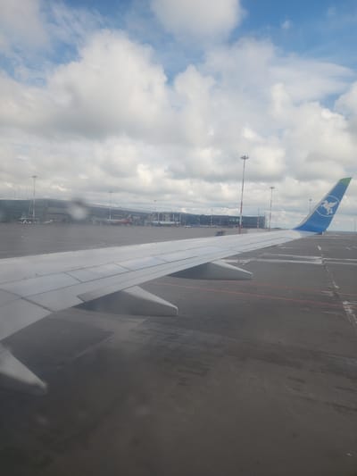 The image depicts a view from an airplane window at the airport in Stolbishche, Russia. Visible is the wing of an aircraft, with a blue tail fin adorned with a white pelican logo. Beyond the wing, an airport apron is observable, featuring tarmac, various airport service vehicles, and terminal buildings. The sky is partially cloudy.