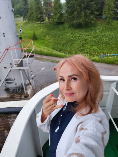 In the city of Belousovo, Russia, a woman with reddish-blonde hair is taking a selfie on a white railing, possibly on a ship or observation deck. She is wearing a white cardigan with a tan pattern over a dark blue shirt. Her right hand is raised, with fingernails painted red, and she is making a gesture with her thumb and index finger. In the background, to the left, a large grey cylindrical structure with red and white stairs is visible. Beyond that, a lush green hillside covered in grass and scattered trees slopes upwards. The ground around the structure appears wet, suggesting recent rain.