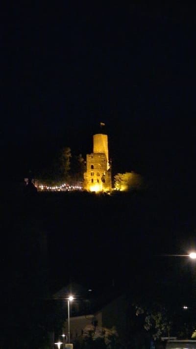 The image depicts a nighttime view of the Strahlenburg castle in Schriesheim, Germany. The stone castle is illuminated with warm, yellow lights, highlighting its weathered texture and architectural details. A tall, rectangular tower stands prominently, topped with a flag. Below the castle, a string of decorative lights is visible, suggesting a festive event. The castle is situated on a hill, with dark, shadowy areas around its base, indicating the surrounding landscape. In the foreground, streetlights cast a glow on a dark, unidentifiable area, with hints of buildings and foliage visible. The sky is completely dark, with no stars or moon discernible. The overall impression is one of a historical landmark illuminated for a special occasion against a backdrop of the night.
