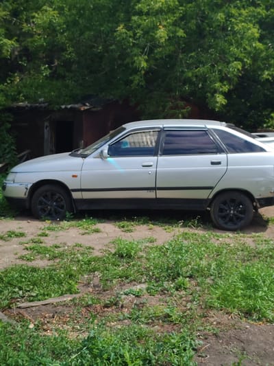 A silver, four-door sedan is parked on a dirt and grass lot. The car is seen from its left side, with the front of the vehicle facing left. The car has tinted windows and black rims. In the background, there are large green trees and a dark, weathered structure with a corrugated metal roof. The ground in front of the car is a mix of dirt and green grass.