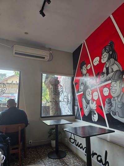This is an indoor scene of a cafe or coffee shop in Klapanunggal, Indonesia. The focus is on a wall with a prominent mural and several tables. The mural depicts several cartoon-like figures in conversation bubbles, rendered in black, white, and red. To the left of the mural, there are two windows showing an outdoor scene with trees and buildings. One window has a sticker with text and graphics that appear to be related to an educational institution. Inside the cafe, a person is seated at a wooden chair, facing away from the camera, looking towards the window. There are two square black tables with pedestal bases in the foreground. One table is closer to the camera, and the other is further back. An air conditioning unit is visible on the upper wall. The floor is tiled with small, rounded stones. The overall atmosphere is casual and seems to be a place for relaxation or socializing.
