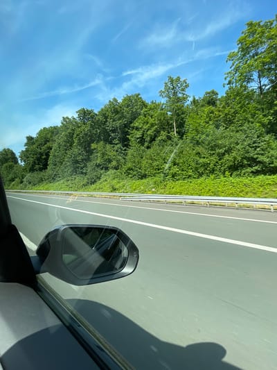 This is a photograph taken from inside a moving vehicle, likely on an autobahn, looking out the side window. The perspective is from the driver's or front passenger's side, showing the side mirror and part of the car's interior. The road ahead is a multi-lane highway with a white line marking the lane. To the right of the road, there is a dense forest with lush green trees and undergrowth. A silver guardrail separates the road from the vegetation. The sky is a clear blue with some wispy white clouds. The overall scene depicts a journey through a scenic, tree-lined highway, typical of areas around Langenfeld (Rheinland), Germany, which is known for its green spaces and Autobahn connectivity. No people are visible, but the presence of a vehicle indicates travel. The lighting suggests it is daytime, likely a sunny afternoon.
