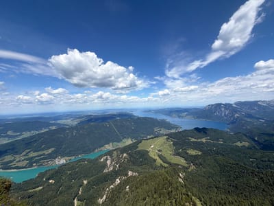 This is a high-angle aerial photograph of the St. Wolfgang im Salzkammergut region of Austria on a clear day with scattered clouds. In the foreground, a steep, tree-covered mountain slope dominates the lower portion of the frame. A rocky outcrop with sparse vegetation is visible on the far left.
Further into the midground, a vibrant turquoise lake stretches horizontally across the image, bordered by a dense green forest. A small village with buildings and white structures is nestled along the lake's edge. Beyond this lake, a larger, deep blue lake extends into the distance, with a long, narrow peninsula jutting out from its right side.
In the background, a series of rolling, tree-covered hills and mountains recede towards the horizon. The far distance is a hazy blue, suggesting a vast expanse of land and water under the bright sky. The sky itself is a brilliant blue, punctuated by large, fluffy white cumulus clouds and thinner, wispy cirrus clouds. There are no people or animals visible in the image. The overall scene depicts a serene mountain and lake landscape.