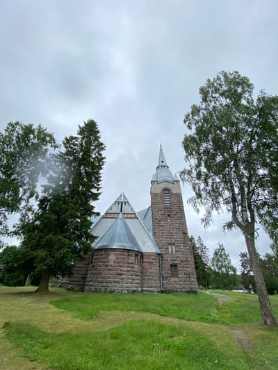 An exterior, low-angle, eye-level shot captures a stone church with a steeple and a peaked roof, set against a cloudy, overcast sky. The church's facade is constructed from rough-hewn, reddish-brown stones. A tall, dark green evergreen tree is positioned to the left of the church, and a lighter-barked tree with green foliage stands to the right, partially obscuring the view of the church's right side. A path winds through a grassy area in front of the church, which slopes upwards towards the building. The scene is located in the city of Melnikovo, Russia.