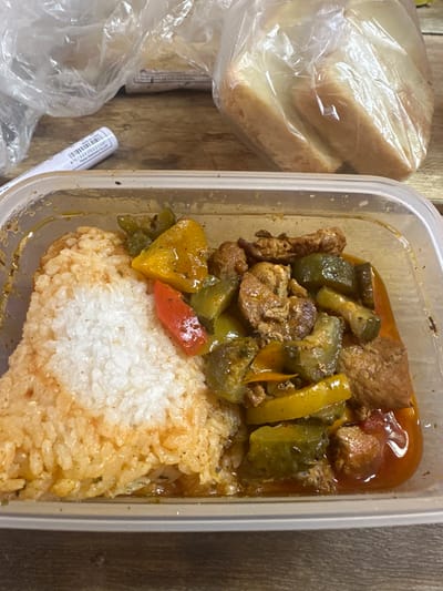 A to-go container of food sits on a wooden surface. The container holds rice and a dish with meat and vegetables. In the background, there are plastic bags with bread. There is no information to indicate this is in the city of Чикшино, Russia.