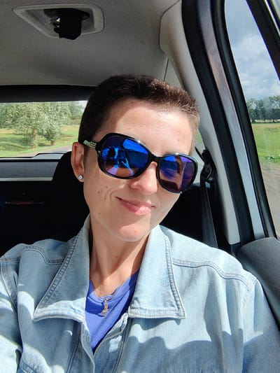 A medium shot photograph shows a woman in a car, looking at the camera. She has short brown hair, wears large dark sunglasses with blue lenses, and a light blue denim jacket over a purple t-shirt. A small, gold necklace with a charm is visible under the collar of the denim jacket. She has a light-skinned complexion with a few freckles and is smiling faintly. The background shows a glimpse of green trees and a blue sky through the car window. The interior of the car is visible, including a black seat and a grey headliner. The photograph appears to have been taken on a sunny day. The image location is outside of Сваткаўскі сельскі Савет, Belarus.