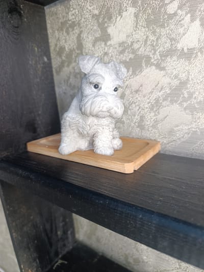 A white, textured figurine of a schnauzer dog sits on a light-colored wooden tray. The tray is placed on a dark wooden shelf. Behind the figurine is a textured, light-colored wall. The image appears to be an indoor scene, possibly a home or shop display. There is no indication of the city of Nikopol, Ukraine, in the image.
