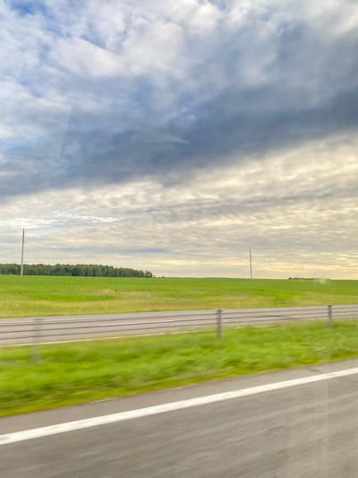 The image shows a wide, open green field bordering a road. The sky is filled with dramatic, textured clouds in shades of white, gray, and blue. The field is lush and appears to be a cultivated crop, with a line of trees forming a dark green border in the distance. Two solitary utility poles are visible in the field, standing against the horizon. The road in the foreground is paved with asphalt and has white lane markings, indicating it is a highway or a major road. A metal guardrail separates the road from the field. The image has a sense of motion, suggesting it was taken from a moving vehicle. The overall scene is one of rural landscape under a dynamic sky. The location is specified as Княжыцкі сельскі Савет, Belarus.