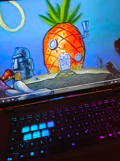 The image shows a laptop screen displaying an episode of Spongebob Squarepants. The screen shows Spongebob's pineapple house, Squidward's house and a view of the seabed. Below the screen is the laptop keyboard with illuminated keys. Some keys are lit in blue and others are lit in pink. The letters 'Q', 'W', 'E', 'A', 'S', 'D', 'Y', 'X', and 'C' are illuminated in blue.
Given the presence of the keyboard with 'å' and 'ö' keys, and the umlaut symbol, the laptop is likely sold in a Nordic country, and not located in Rüsselsheim am Main, Germany.