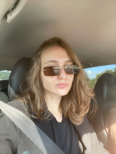 A young woman with shoulder-length, light brown hair is in the driver's seat of a car, wearing sunglasses and a black top under a dark blazer. She is looking at the camera with a neutral expression. The seatbelt is visible across her chest. The interior of the car, including the headrest of the passenger seat, is visible. Outside the car, trees and a blue sky can be seen through the windows. There is no indication of the city of Shud'ya, Russia in this image.