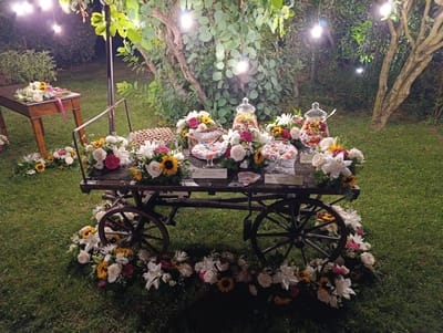 The image depicts an outdoor evening setup, likely for an event, in the city of Scandiano, Italy. A rustic wooden cart, adorned with abundant floral arrangements featuring white lilies, roses, sunflowers, and pink accents, serves as a prominent display. On the cart's surface, glass jars filled with colorful candies and smaller glass bowls also containing candies are arranged among the flowers. Small labels with Italian text, such as "Ginevrine Senza Glutine," are visible. To the left, a separate wooden table holds additional floral arrangements. The scene is illuminated by several hanging lightbulbs, casting a warm glow on the surroundings, which consist of green grass and trees with lush foliage. The overall impression is one of a charming and festive outdoor display.