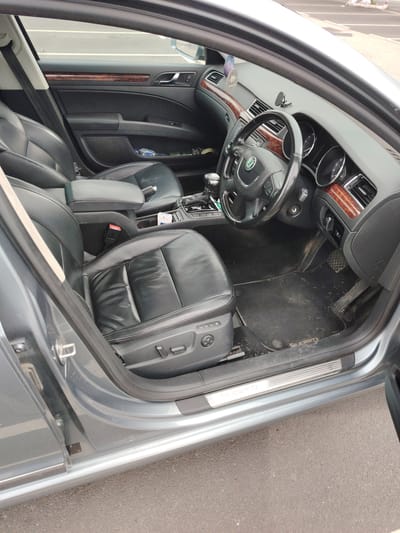 The image displays the interior of a grey Skoda Superb, viewed from the open driver's side door. The vehicle features black leather seating, with the driver's seat showing electric adjustment controls and creasing on the upholstery. The dashboard and door panels are accented with dark wood-grain trim. A black multi-function steering wheel with the Skoda emblem is visible. A purple air freshener labeled "89 aromatic" hangs from the rearview mirror area, and a black phone mount is affixed to the dashboard. The center console contains an automatic gear selector, a central armrest, and a white box, possibly a cigarette packet, in a cup holder. The black floor mats are dirty with accumulated dust and debris, with the "SUPERB" logo visible on the driver's mat. A plastic water bottle is present in the driver's door storage pocket. The scene depicts the vehicle parked on asphalt pavement with painted lines visible in the background, located in Peterborough, United Kingdom.