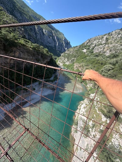 The image presents a first-person elevated view from a rustic suspension bridge overlooking a river canyon under a clear blue sky with minimal white clouds. A prominent multi-strand steel cable, exhibiting significant rust, spans horizontally across the upper portion of the frame. Below it, rugged mountainsides rise steeply, covered in dense green deciduous and coniferous vegetation interspersed with exposed grey rock formations.

At the base of the canyon, a clear, turquoise-blue river flows, revealing its pebbled and rocky bed through the water. The river varies in depth, indicated by lighter and darker shades of blue-green, and is bordered by rocky banks with some sparse vegetation. In the mid-right foreground, a tanned human right arm, featuring visible hair, extends into the frame, with the hand grasping a section of the bridge's rusted metal mesh railing. This mesh, constructed from square-patterned heavy gauge wire or thin rebar, displays extensive reddish-brown oxidation. The bridge structure itself appears weathered. The scene is set in the natural, mountainous environment of Unknown, Montenegro.