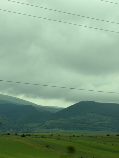 A landscape photograph, likely taken in the city of Mrgvali Chala, Georgia, features a view of rolling hills and overcast skies. The foreground contains a series of power lines stretching vertically across the frame. To the right, the landscape transitions from mountainous terrain covered with trees to open, green fields. The sky is predominantly gray, suggesting cloudy conditions. The image is oriented sideways.