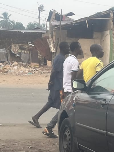 A car is parked along a dirt road in front of a rundown neighborhood. Three men stand near the car. One man wears a yellow shirt, another a white shirt, and the third a dark shirt. The environment is unkempt with weathered buildings and debris. Power lines are visible in the background. A coconut tree is also seen in the bottom left.