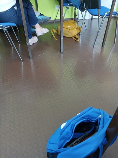 This indoor image depicts a scene within what appears to be a classroom, lecture hall, or study area in Kaiserslautern, Germany. The floor is covered with brown-grey rubberized tiles featuring a textured pattern of raised circular studs.
On the left side of the frame, a person is seated at a desk. Only their lower body is visible, wearing blue jeans and two distinct shoes: a dark athletic shoe on one foot and a light beige platform shoe or boot on the other. Underneath the chair and desk, a bright yellow backpack rests on the floor.
A prominent blue backpack is situated on the floor in the foreground on the right. Its main compartment is open, revealing contents that include a magazine or textbook with partial text visible, appearing to read "TEPAT" or "TEPATS."
The setting is furnished with multiple metal-framed chairs, some with blue seats, and corresponding tables. A bright lime green wall is visible in the background on the left, featuring a white electrical outlet. The lighting suggests an indoor environment, casting shadows of the furniture onto the patterned floor. No specific time of day or outdoor weather is discernible. The overall impression is one of a functional, possibly academic, interior space.
