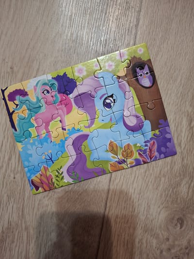 The image shows a partially completed jigsaw puzzle depicting two cartoon ponies, placed on a light-colored wooden surface. The pony at the top is light blue with a purple mane and tail, and the pony at the bottom is pink with turquoise hair. An owl is visible in the top left corner. Floral and foliage designs surround the ponies. There is no evidence of the city of Ozyorny, Russia.