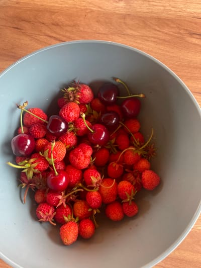 A light grey-blue ceramic bowl, partially visible on a wooden surface, contains a dense cluster of fresh fruits. The majority of the fruits are small, red wild strawberries or similar garden strawberries, displaying a textured surface and some with attached green calyxes. Interspersed among the strawberries are approximately ten dark red, almost black, cherries, each with a slender green stem. The fruits are tightly packed, filling the lower portion of the bowl. The wooden surface beneath the bowl exhibits a distinct grain pattern in shades of light brown and orange-brown. The scene is illuminated by ambient light, creating soft shadows within the bowl and highlighting the natural textures of the fruits and wood. The photograph depicts these items in Petrivka, Ukraine. No individuals or actions are depicted.