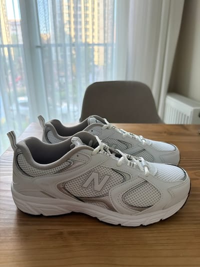 The image shows a pair of white New Balance athletic shoes with grey accents, placed on a wooden table. The shoes are positioned side-by-side, with the left shoe slightly closer to the camera. The background features sheer white curtains, a blurred view of buildings, and a chair. The lighting suggests an indoor setting, possibly a room with a window. The scene appears to be located in Zeytinburnu, Turkey, as indicated by the presence of buildings and curtains commonly found in residential areas there.