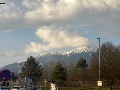 The scene depicts a wide outdoor view in Radovljica, Slovenia, under a partly cloudy sky with prominent white and grey cumulus clouds against patches of blue. In the background, a range of snow-capped mountains with visible rock faces and dark evergreen tree lines dominates the horizon. The midground features a dense line of bare deciduous trees, interspersed with several dark green coniferous trees, separated from the foreground by a wire fence. A lattice-structure power line tower is visible on the far left of the midground. The foreground reveals a paved area, likely a parking lot or road shoulder, occupied by several parked passenger vehicles, including a white car and a dark-colored car. Prominent street furniture and signs are present: on the left, a large circular blue sign with a red diagonal line through a white rectangle (indicating "no stopping" or similar restriction), stacked above a smaller white square sign depicting a truck with a camera icon. To the right, a tall, slender, dark grey street lamp stands erect. Partially visible is a structure resembling a bus stop or kiosk, featuring advertisements, and a yellow triangular warning sign with a black border and red symbol below the street lamp. No individuals are visible.