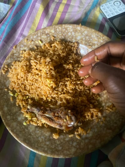 An overhead view depicts a meal in an indoor setting within Idiroko, Nigeria. A dark-skinned hand is actively engaged in consuming jollof rice from a round, speckled brown and beige ceramic plate. The rice exhibits a reddish-orange hue with interspersed lighter yellow grains, and a bone fragment, likely from a poultry part, rests on the plate. The hand's fingers are slightly glossy, indicating contact with the food. The plate and hand are positioned on a striped fabric surface featuring vertical bands of purple, yellow, and blue. In the upper right quadrant of the scene, a white and black feature phone with a visible keypad is resting on the same striped fabric.
