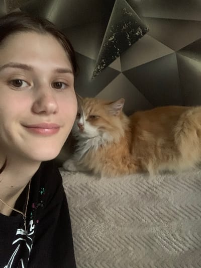A young person with brown hair, looking at the camera, is in the foreground of the image. They are smiling and have a necklace on. To their right is a fluffy orange and white cat, also looking towards the camera. The cat appears to be lying down. They are both on a textured, light-colored surface. The background is a dark, geometric patterned wall. The scene appears to be indoors, likely in a home environment.