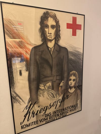 This is a poster featuring a stark, charcoal-style illustration of two figures, an adult and a child, seemingly in distress. The adult, with long, dark hair and a somber expression, holds the child who rests their head on the adult's shoulder, their face also conveying a sense of weariness or sadness. Both figures are depicted in dark clothing, with the adult wearing what appears to be a jacket. Behind them, the background is rendered in rough, abstract strokes, suggesting a chaotic or somber environment.
Prominently displayed in the upper left corner is a large red cross, a symbol universally associated with the Red Cross and humanitarian aid. To the right of the figures, bold, black text is inscribed vertically. The main title reads "Kriegsopfer," which translates to "War Victim" or "War Sufferer" in German. Below this, it further elaborates: "DAS INTERNATIONALE KOMITEE VOM ROTEN KREUZ HILFT!" (The International Committee of the Red Cross Helps!). Additional text provides a postal check number and location: "POSTCHEK I. 777 GENF," indicating Geneva, Switzerland, as the contact point.
In the lower left portion of the poster, there is some red and black text in a different script, partially obscured and difficult to decipher fully, but it seems to contain phrases like "Hilfe" (Help) and possibly a date or reference. The overall tone of the poster is one of somber urgency, aiming to evoke empathy and solicit support for victims of war through the Red Cross. The style of the illustration and the text suggest it is a historical piece, likely from a period of significant conflict. The poster is framed and mounted on a white wall, possibly in a gallery or museum setting.