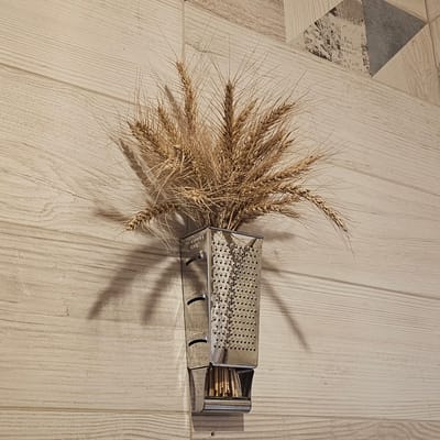 A cluster of dried wheat stalks is arranged inside a metal grater. The grater is positioned on a light-colored wooden surface, possibly a floor or table, with visible wood grain. To the right of the grater, the grater's drawer is pulled out, revealing a bundle of matchsticks. The scene is illuminated, casting shadows from the wheat and the grater onto the wooden surface. The background shows a section of a wall with a geometric pattern composed of wood-toned and grey triangular shapes, some with distressed text. The overall impression is still life, likely an indoor setting. No people are visible. The time of day and weather are not discernible. The location context is Vybolovo, Russia, but there are no specific visual cues within the image to confirm this. The grater has some faint text visible on its side, but it is illegible in the image.