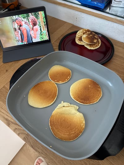 A bird's-eye-view shot shows a tablet displaying a scene with two people outdoors, a plate with several pancakes, and a square gray griddle with four pancakes in various stages of being cooked. The location is likely indoors on a wooden surface, possibly a kitchen counter or table. The scene appears to be in a residential setting. The pancakes on the plate are browned and stacked. The pancakes on the griddle are golden brown and appear to be close to being finished. The tablet is black and has a gray case. No information indicates this is in San Rafael, United States of America.