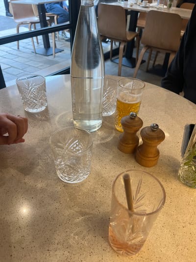 The image displays a tabletop scene within a restaurant or cafe, likely located in Aix-en-Provence, France.
**Subjects and Objects:**
On a light-colored, speckled table surface, a tall glass bottle, approximately half-filled with clear water, stands centrally. To its left, an empty, lowball-style glass with an etched pattern is visible. Another similar empty glass is positioned further down and to the left. To the right of the water bottle, a pilsner glass containing a golden-yellow liquid, consistent with beer, is present. Adjacent to the beer glass are two light-brown, wooden pepper and salt shakers with silver tops. In the lower right foreground, a partially consumed drink in an etched glass, showing a small amount of pinkish liquid and containing a light-colored straw, is seen.
**People and Actions:**
A person's left hand, adorned with a ring on the fourth finger, is visible in the lower-left corner, resting near one of the empty glasses. A partial view of a person's torso, wearing a dark garment, is visible on the right side of the table, suggesting a diner.
**Environment:**
The background reveals the interior of a dining establishment with light-colored wooden chairs and dark-legged tables. Beyond the interior, a large window or glass partition provides a view of an outdoor paved area, suggesting a street or sidewalk, with additional seating visible. The overall lighting is bright, casting some shadows on the table surface.
