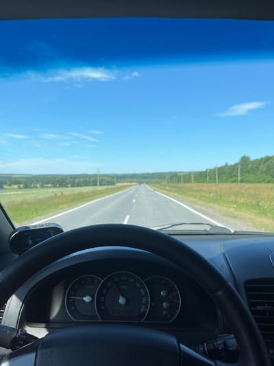 The image captures a view from inside a car driving on a straight, paved road through a rural landscape. The sky is a clear, vibrant blue with scattered white clouds. The road ahead extends into the distance, bordered by grassy fields and trees on either side. Utility poles are visible along the right side of the road. The car's steering wheel and dashboard are prominent in the foreground, with the speedometer showing a speed of approximately 70 km/h. A small digital display unit is mounted on the dashboard. The overall scene suggests a bright, sunny day and a journey through the countryside. The location is identified as being in the city of Kukshumy, Russia.
