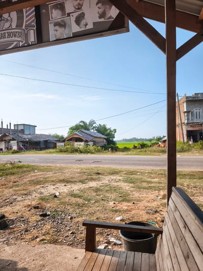 The image captures a scene in Blangmajron, Indonesia, focusing on a roadside view from a covered wooden structure. In the foreground, a wooden bench is partially visible, suggesting a waiting area. To the left, a black bucket sits on the ground next to a wooden beam. Above, a sign displays images of men's hairstyles, indicating the establishment is likely a barbershop. The middle ground features a paved road with dry, sparse vegetation on either side. Beyond the road, several modest houses with thatched or tin roofs are clustered together, some appearing weathered. Further back, lush green rice paddies are visible, and a small herd of cattle grazes in the field. A multi-story building with an awning labeled "AC A" stands on the right side of the road, with a few people near its entrance. The sky is clear and blue, with a few wispy clouds. The overall impression is of a rural or semi-rural setting with a mix of residential, commercial, and agricultural elements.