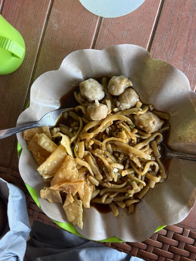 A close-up, overhead shot shows a bowl of noodles with meatballs and fried wontons served on a wooden table. The noodles are coated in a dark sauce and mixed with chopped green onions and red pepper flakes. Four meatballs are visible on top of the noodles, and several pieces of crispy fried wontons are scattered around the edges of the bowl. A spoon rests on the left side of the bowl, and another is partially visible on the right. A bright green plastic object, possibly a condiment dispenser, is in the upper left corner of the image. A light blue cloth or napkin is visible in the lower left corner, and a brown woven surface is in the lower right. The scene appears to be an outdoor dining setting, potentially at a street food stall in Welado, Indonesia.