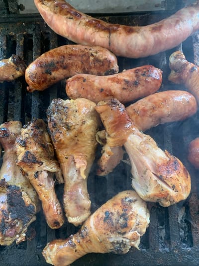 The image displays a close-up, top-down view of various meats actively cooking on a dark, metal grate barbecue grill in Lauro de Freitas, Brazil. The grill surface is visibly worn and appears seasoned with grease and char residue. Multiple pieces of meat are arranged across the grates. In the upper portion, a large, light-pink sausage with some browned marks extends horizontally. Below it, four shorter, thicker sausages show more extensive browning and localized charring. The lower half of the frame is dominated by approximately seven chicken drumsticks and possibly other chicken pieces, exhibiting golden-brown skin with distinct grill marks and darker, charred sections. Wisps of white smoke are visible rising from between the grates and around the cooking meat, indicating an active heat source. No individuals are present in the frame.