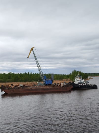 The image shows a barge with a crane on it docked next to a tugboat on a body of water. The barge is loaded with a pile of sand and debris. The crane is gray and blue with a yellow boom. The tugboat is black and white with the name "ПРИБОЙ" visible on its side. The background consists of a shoreline with trees and reeds under a cloudy sky. The location is identified as Ozernoe Ust'e, Russia.