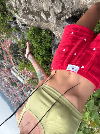 A person is positioned on a rocky incline overlooking a densely populated urban area with red-tiled roofs, likely in Tam Thanh Ward, Vietnam. The person is wearing a light green ribbed tank top and red shorts with white heart patterns. The image is taken from a high angle, looking down at the scene.

The person's torso is visible, with a subtle hint of a belly button. A tattoo is visible on their left arm, featuring stylized designs including what appears to be a palm leaf. The person is also wearing a bracelet with colorful beads on their left wrist. A thin black braid extends from the person's hair, partially across their chest. A delicate gold necklace is also visible.

The surrounding environment is characterized by rough, grey rocks and lush green foliage. The urban landscape below suggests a daytime scene, with no clear indicators of weather. The overall impression is one of a person enjoying a scenic overlook.

A small white tag is attached to the person's shorts. Visible text on the tag includes "EZWOOL" in large letters, with smaller text above and below. "REASHION THE FUTURE" is also visible.