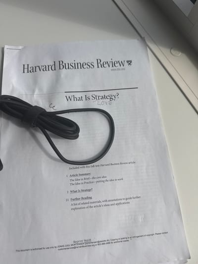 The image shows a close-up of a document from the Harvard Business Review. The document appears to be a printout of an article titled "What Is Strategy?". There is a black power cord coiled and secured with a velcro strap lying on top of the document. Handwritten text "LOVE" is visible on the document, near the title. The bottom of the document contains reprint information and a copyright notice. The scene is indoors, with the document placed on a light-colored surface, possibly a desk. The lighting suggests it is daytime. There is no indication of the city of Aarhus, Denmark, in the image.