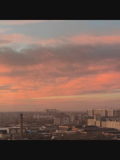 This is a wide-angle aerial shot of a cityscape under a dramatic sunset. The sky is filled with clouds in shades of pink, orange, and grey, suggesting a warm, possibly humid, atmosphere. The sun is likely low on the horizon, casting a warm glow over the scene.
The foreground is dominated by industrial buildings and a tall, brick chimney stack. Further in the background, residential buildings and taller structures, some appearing to be apartment blocks, are visible. Cranes can be seen scattered across the urban landscape, indicating ongoing construction. The overall impression is that of a functional, lived-in city.
There is no visible text in the image. The scene evokes a sense of quiet observation, perhaps a moment of calm at the end of the day. The interplay of warm light from the sunset and the muted tones of the urban environment creates a visually striking contrast. The location context places this in Saint-Août, France.