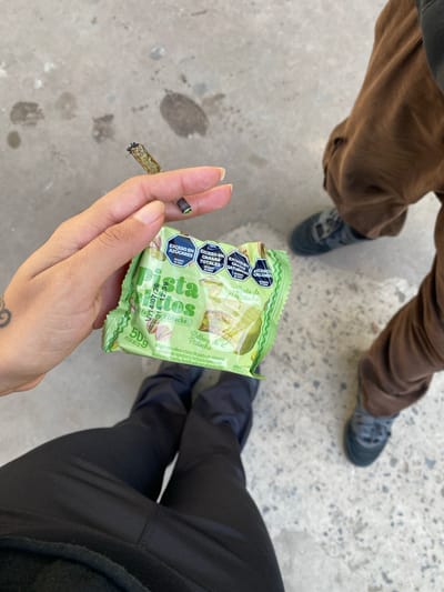 The scene presents an overhead shot of a person's hand holding a partially smoked joint, along with a green package of "Alfajor Relleno de Pistacho" candy. The packaging contains nutritional information, highlighting excess sugars, total fats, saturated fats, and calories. The setting appears to be outdoors on a concrete surface, with two other individuals standing nearby; one in brown corduroy pants and gray shoes, and another in black pants and dark shoes. There's no verifiable indication that the location is in Monte Grande, Argentina, other than the text on the candy wrapper indicating that it is an "Industria Argentina" product.