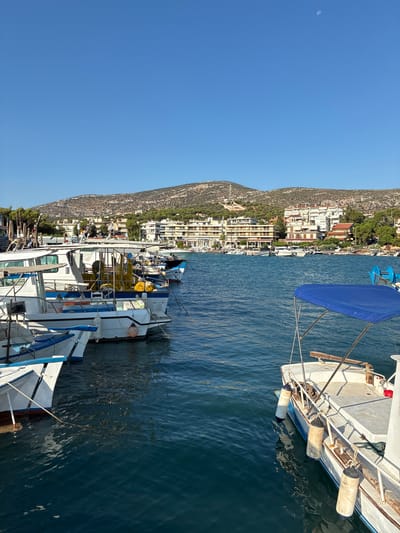 The image depicts a clear, sunny day in the harbor of Limenas Markopoulou, Greece. The foreground is dominated by several docked boats, primarily white with blue trim, some of which appear to be fishing vessels equipped with yellow netting. One boat on the right has a blue canopy. The water is a deep blue, reflecting the clear sky. In the background, a row of buildings, including hotels and houses, line the shore. Behind the buildings, a tree-covered hill rises, with the peak appearing somewhat arid and rocky. The sky is a vibrant, cloudless blue, with a faint crescent moon visible near the top left. No people are visible in the image.