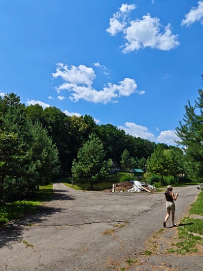 A woman wearing a dark top, tan shorts, and a baseball cap walks away from the camera on a cracked asphalt path. She holds a light-colored cup in her right hand. The path curves to the right and continues into a grassy area with a line of trees along its edge. To the left of the path, more asphalt is visible, leading to a split in the path. Beyond the path is a dense forest with a clear blue sky and scattered white clouds above. In the background, there are a few small structures, including a green-roofed gazebo and a pile of debris. The overall scene depicts an outdoor setting on a sunny day.