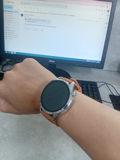 The image shows a close-up of a person's left forearm and wrist, wearing a smartwatch with a brown strap. The watch face is dark and circular. In the background, a computer monitor displays a Gmail inbox with an email open. The monitor is turned on and shows a web browser window. A black keyboard is visible below the monitor. The scene appears to be an indoor setting, possibly an office or home, with a neutral-colored surface in the foreground. The image quality is slightly blurry, particularly in the background. There are no discernible landmarks or features in the image that would indicate the location as Tokmok City, Kyrgyzstan.