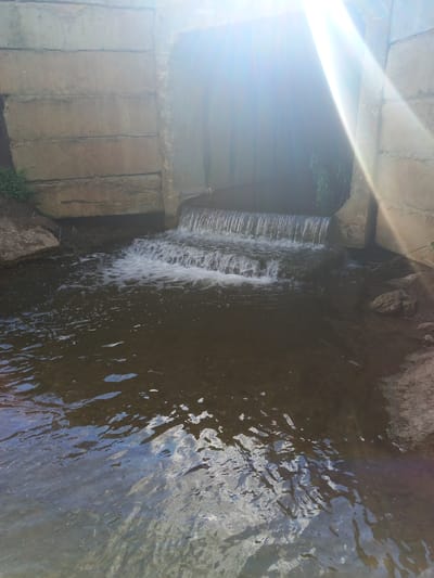 The image shows a small waterfall in a narrow channel, likely a stream or culvert, surrounded by concrete and natural rock. The water is flowing over a series of small drops, creating a white foamy cascade. The water itself is dark and appears to have a rocky or muddy bottom visible in some areas. The reflections on the surface of the water suggest a bright, possibly cloudy sky.
On the left side of the frame, there are concrete structures that appear to be part of an embankment or bridge. The concrete is weathered and shows signs of age. A strong sun glare is present in the upper left quadrant of the image, obscuring some details and casting a bright light across the concrete. Hints of green foliage are visible in the background, suggesting a natural or semi-natural outdoor setting.
The perspective is from above and looking down into the channel. The overall scene is one of flowing water in a man-made and natural environment. The time of day appears to be daytime, given the strong sunlight and reflections. There is no text visible in the image. The specific location is Sterlibashevo, Russia.