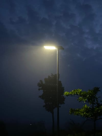 The image, presented rotated 90 degrees counter-clockwise, depicts a nighttime outdoor scene in Amaigbo, Nigeria. A bright, rectangular street lamp is prominently featured on the left side, emitting an intense yellow-white light that extends horizontally into the frame. This powerful illumination creates a strong, diffused halo around the light source, suggesting the presence of mist or humidity in the air.
Above the lamp (which would be the upper-left sky in an upright orientation), the sky is dark and cloudy, characterized by distinct, textured cloud formations against a deep blue-grey backdrop. Below and to the right of the lamp, dark silhouettes of trees are visible, with some patches of green foliage brightly illuminated by the intense light. The far right section of the image displays a dark, reflective surface, likely water, mirroring the bright street light and the indistinct shapes of the surrounding trees.
The scene conveys a quiet, nocturnal ambiance, devoid of human activity or any visible text. The notable details include the nighttime setting, the cloudy weather, and the reflective water surface.