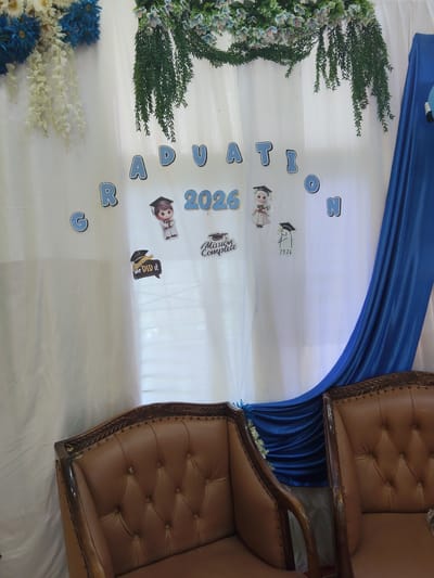 The image depicts a graduation-themed backdrop, likely set up for a photo opportunity. A white curtain serves as the primary background, draped with blue fabric on the right side. Suspended from the top are artificial floral arrangements and greenery. Prominently displayed on the curtain are the words "GRADUATION" and "2026" in blue, block-style letters. Scattered around these words are cartoon-like stickers of individuals wearing graduation caps, with one sticker reading "Mission Complete" and another saying "WE DID IT." A small sticker with "2026" is also visible. In the foreground, two plush brown leather armchairs are positioned, suggesting a seating area for guests. The overall scene suggests a festive occasion, possibly at a school or event venue in Kutalhoksukon, Indonesia. There are no individuals present in the image.
