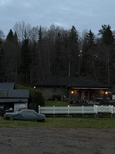 The image depicts an outdoor scene in Jessheim, Norway, at dusk under an overcast, grey sky. In the foreground, a muddy, disturbed ground surface is visible. To the left, a grey car cover conceals a vehicle, exhibiting an elongated form, parked on the muddy terrain adjacent to a dark, single-story outbuilding with a dark roof and a partial view of its contents through an open section.

In the midground, a vibrant green lawn is bordered by a white picket fence, featuring individual, luminous white lights atop several posts. Behind this fence, the main subject is a single-story residence constructed with grey siding and a dark tiled roof. The house facade is illuminated by several active exterior wall lights. A prominent glass door entrance, flanked by additional lighting, leads to a covered outdoor seating area furnished with a table and chairs. An air conditioning unit is affixed to the exterior wall below a window.

The background is dominated by a dense, dark forest consisting of both bare deciduous trees and dark green coniferous trees. Several distant, faint lights are discernible within the tree line. No individuals or discernible activities are present in the scene.